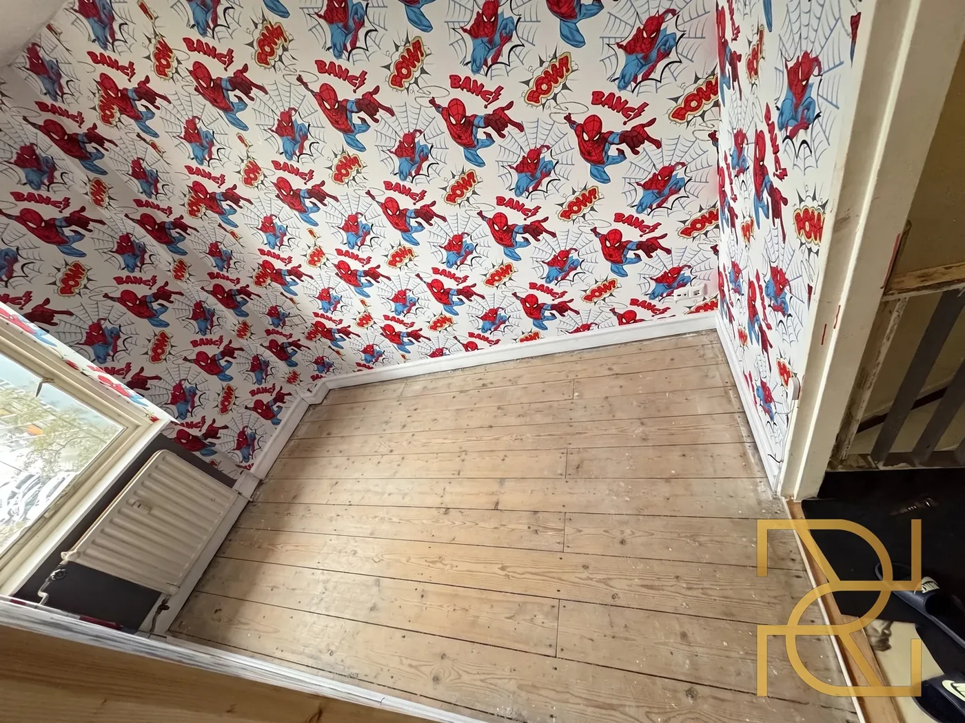 De kamer voor de uitvoering: een kale houten vloer van brede grenen delen onder het Spider-Man-behang